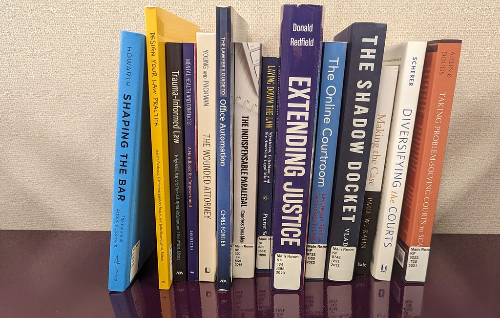 Fourteen books of varying sizes and colors are standing up on a dark purple surface. The spines of the books face the camera so that their titles are visible.