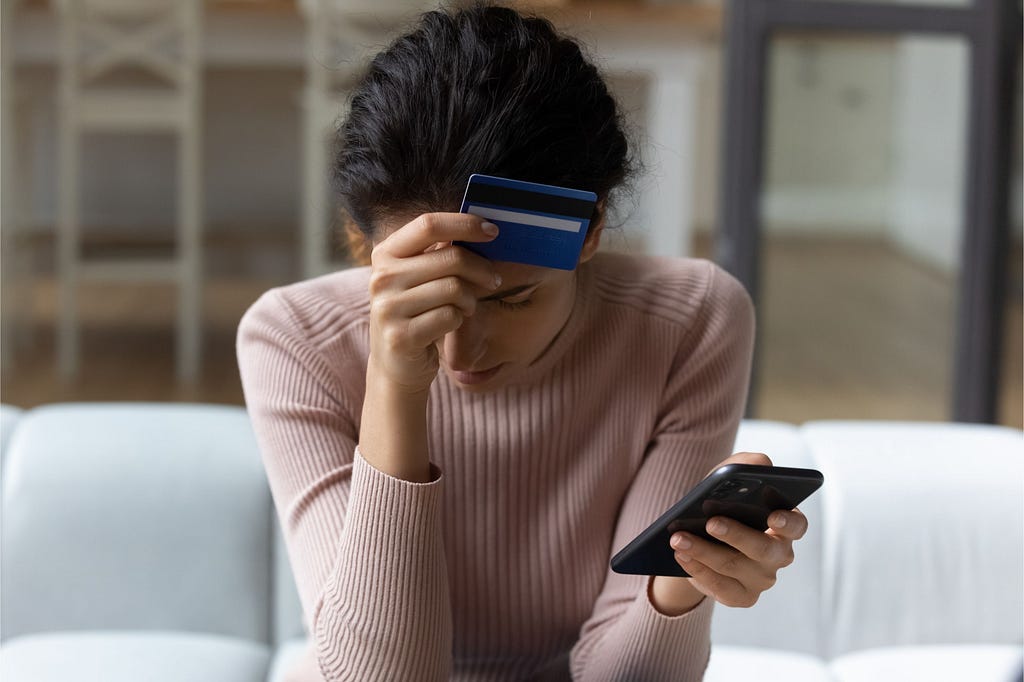 Stressed woman sitting on couch holding credit card and phone.