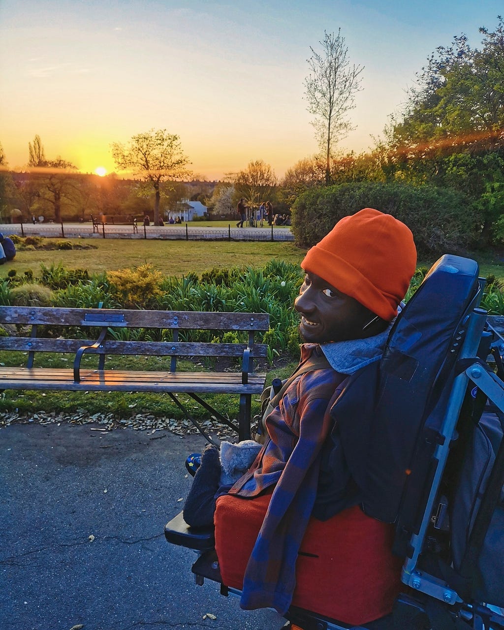 A Black man in his wheelchair in a park facing the sunset.