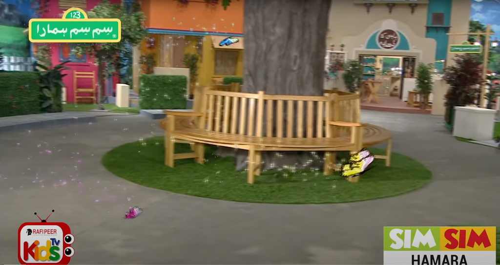 A screen grab of the TV show showing a tree surrounded with benches and three animated butterflies, a large yellow, and two small blue and pink flying