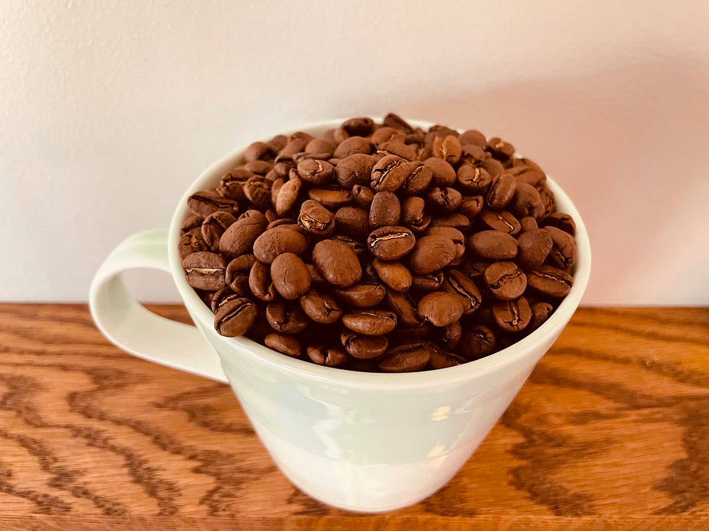 A coffee cup full of coffee beans sits on a stained oak shelf.