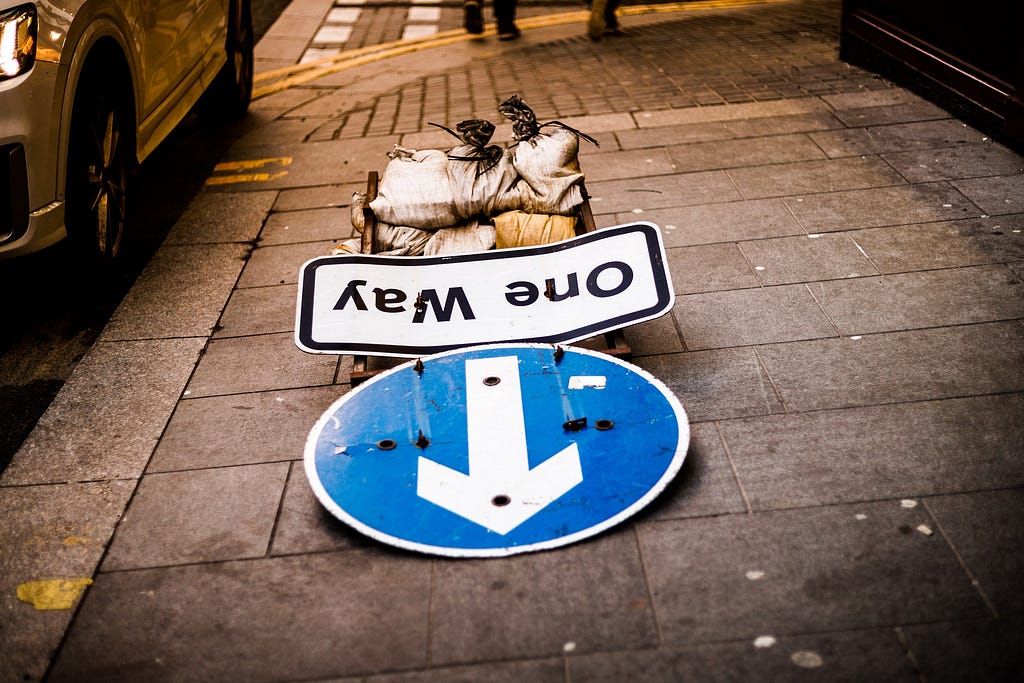 A sign that reads “One Way” is laid horizontally along a sidewalk, having either been taken down or knocked over. Its letters are upside-down from our perspective, and its arrow & intended direction are pointing straight back at the camera. It would appear we are “going the wrong way.”