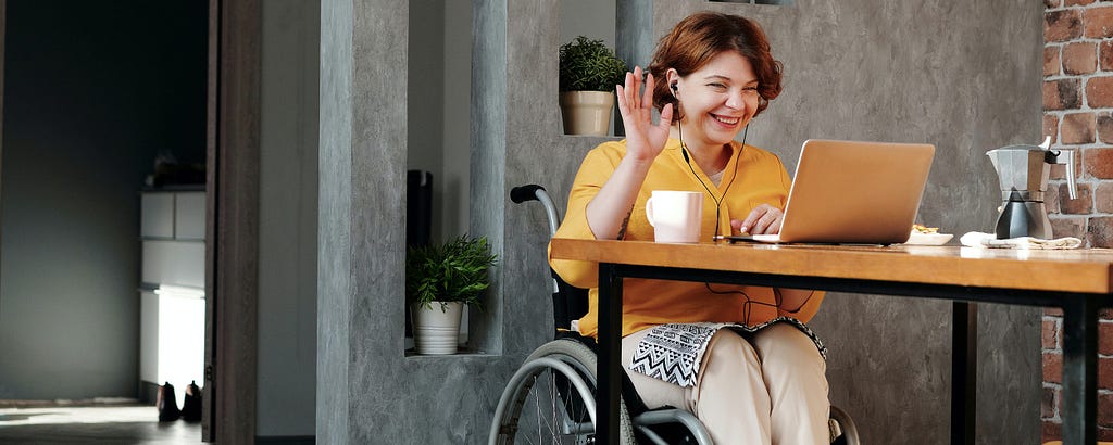 Woman in a wheelchair, smiling and wearing a yellow shirt, sitting at a table with a laptop.