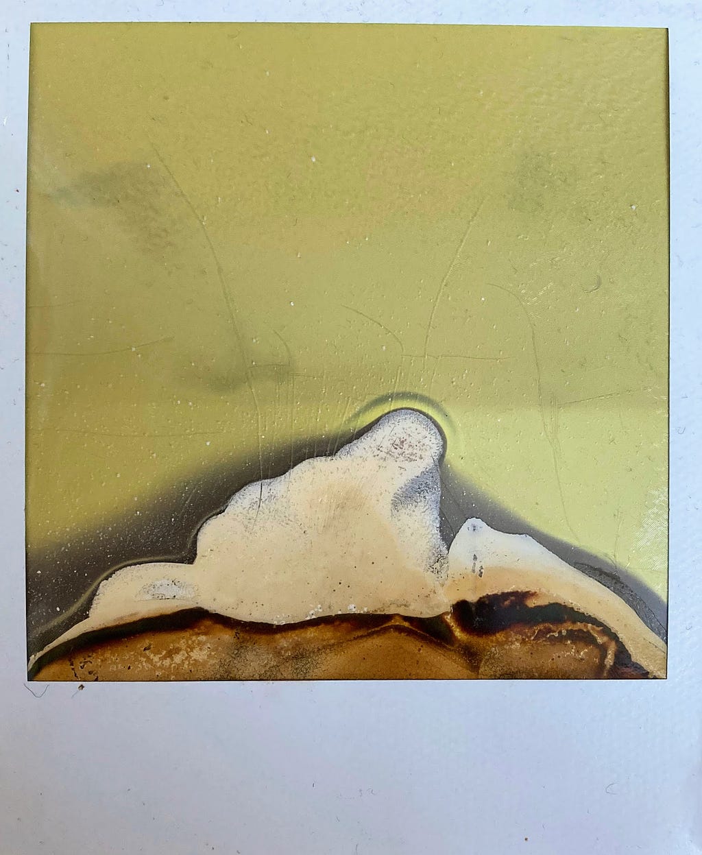 A polaroid photo made with expired film in green-brown sepia tones. The chemicals spread upwards forming a small hill with green-brown and white-ish molten strata. The polaroid itself is old and a bit dusty,and cracked in places.
