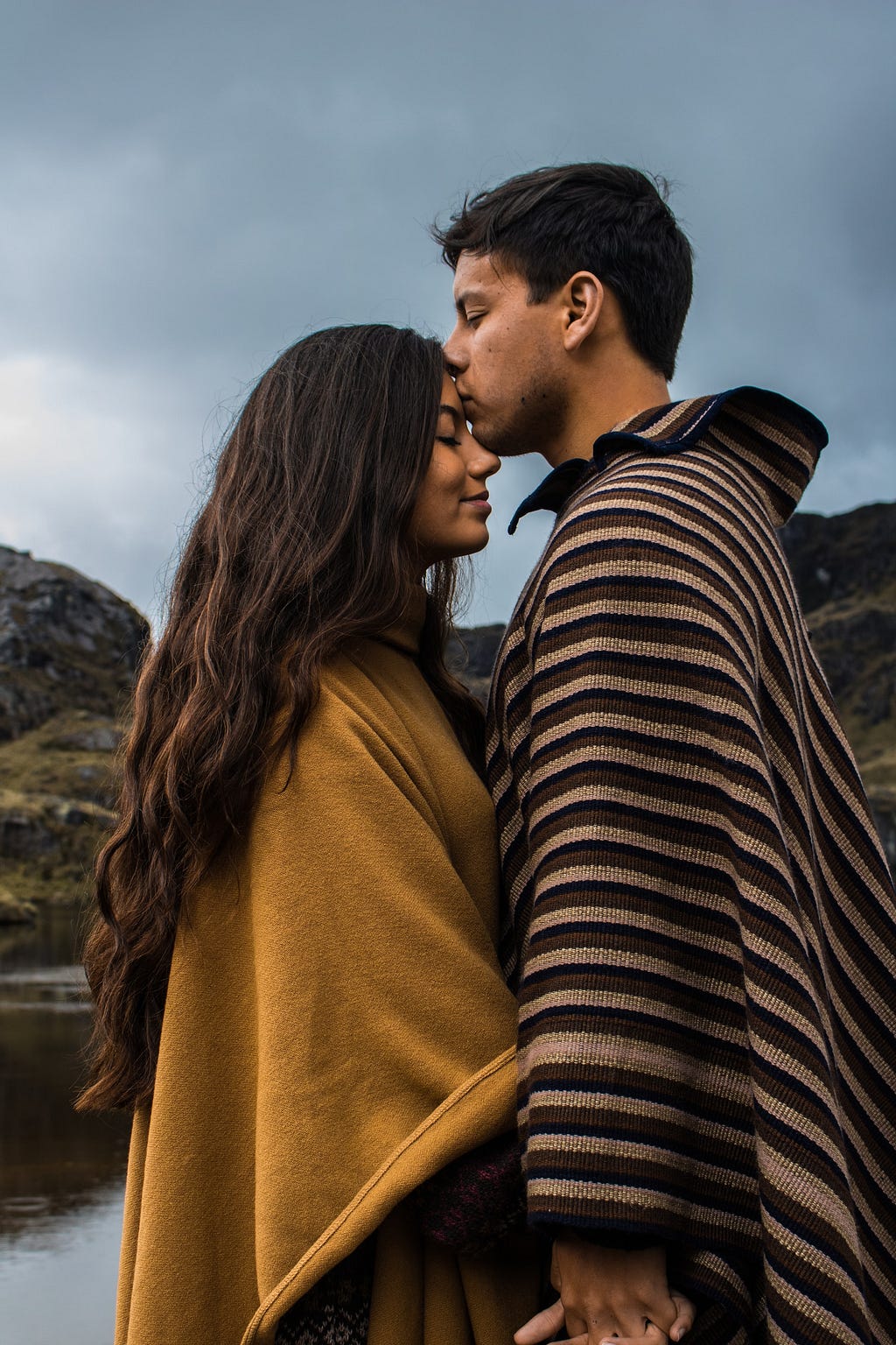 Photo of two young people with brown skin & dark hair, standing close, facing each other with eyes closed. Her face is smiling. She slightly tilts her head up. Her forehead meets his lips as he kisses her. She wears a mustard yellow-brown cloth poncho, and he wears a brown-tone, striped cloth poncho. They hold hands, with their arms down, only slightly visible under the ponchos. The calm water, and rocky, green mountains are in the background, andthe blue sky is spotted with white clouds.