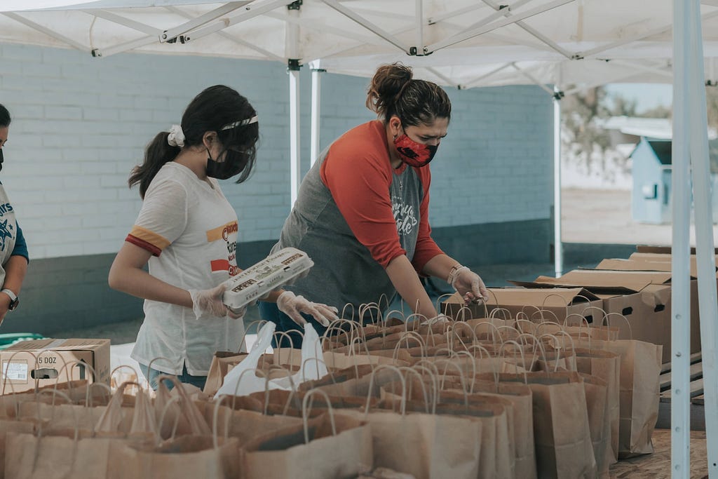Two women in T-shirts in front of a bunch of open paper bags with handles. One holds an egg carton with a dozen eggs, and the other is opening a bag and reaching inside.