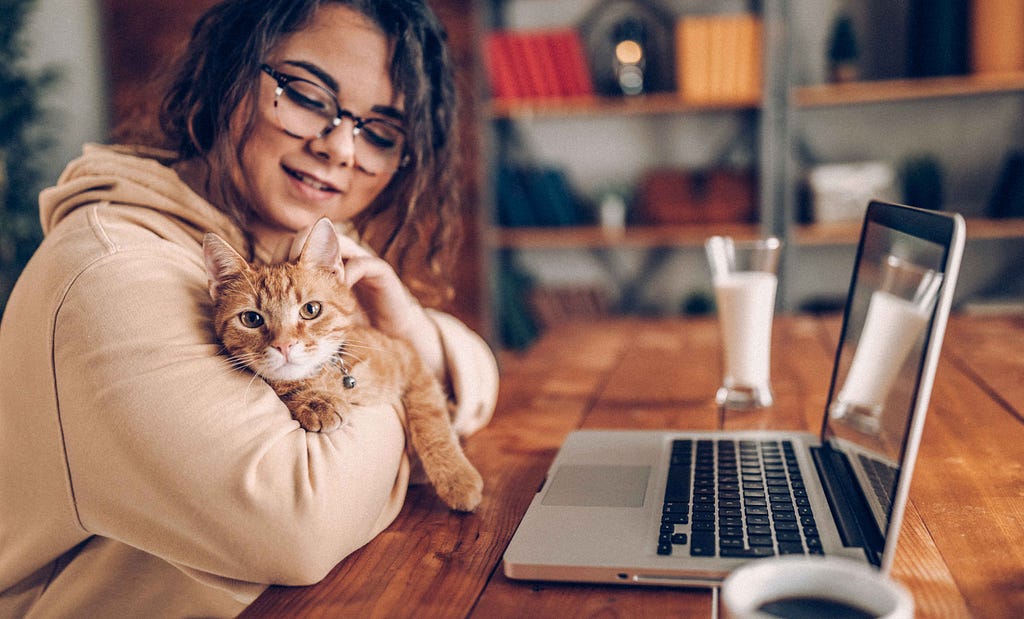 A young woman sitting by the desk working from home with a cat on her arms.