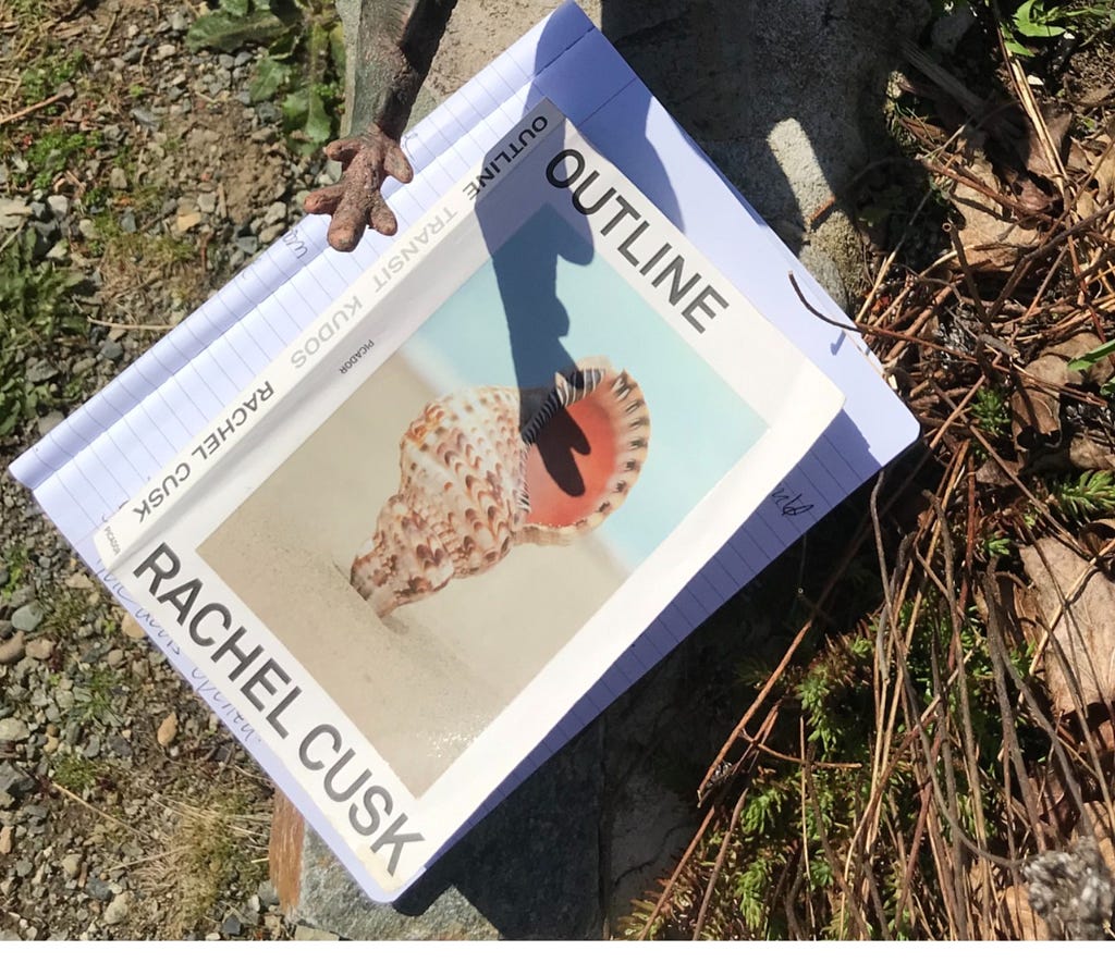 Book cover on dried grass, Outline by Rachel Cusk, with shadow of dinosaur tail over book.