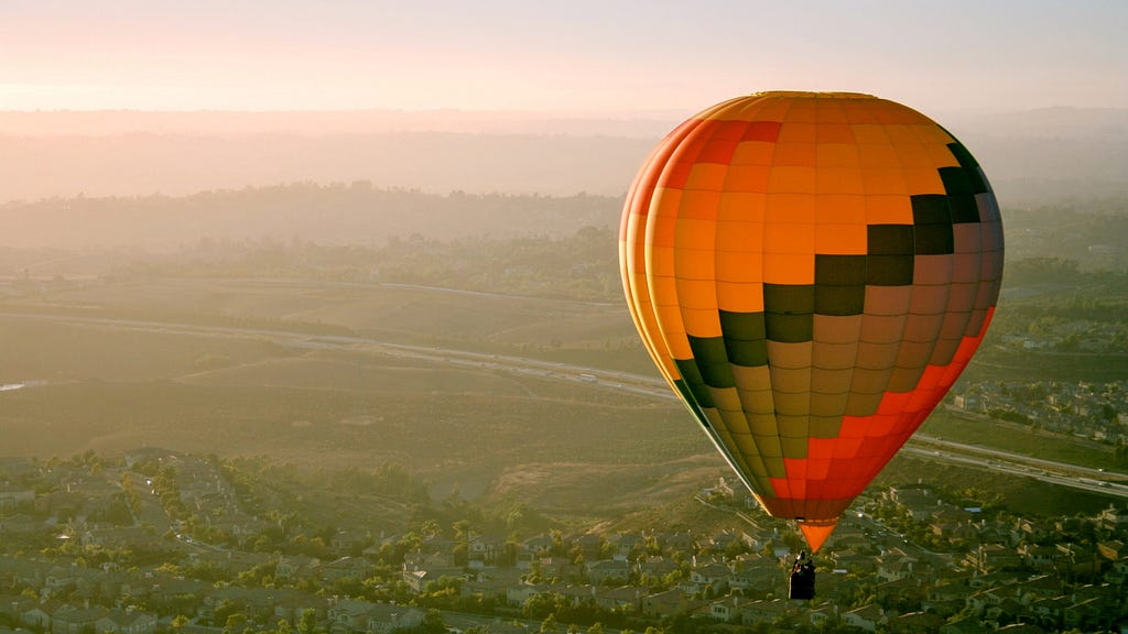 A hot air balloon hovers high over homes and roads with mountains in the distance at sunset.