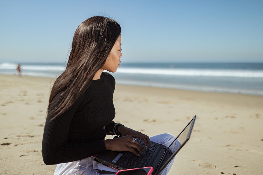 Woman in black long sleeve shirt using black laptop pink phone case working on computer at the beach looking out in the distance remote work