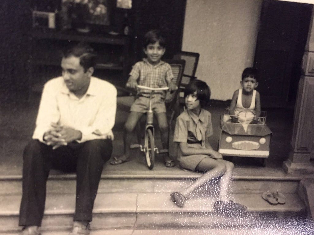 A black-and-white photo shows a man sitting on the steps with a young girl has two young boys also posed for the picture, one on a bicycle and another in a toy pedal car.