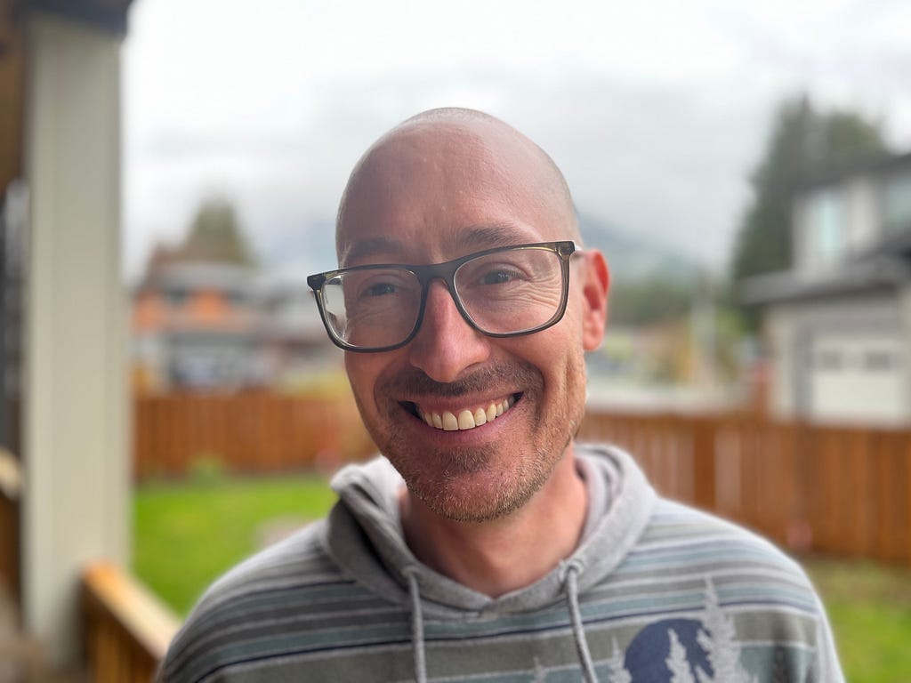 A photo of Andrew with a backyard with grass and a wood fence in the background. He wears a gray striped hoodie, has a shaved head, and wears large black glasses. He is smiling.