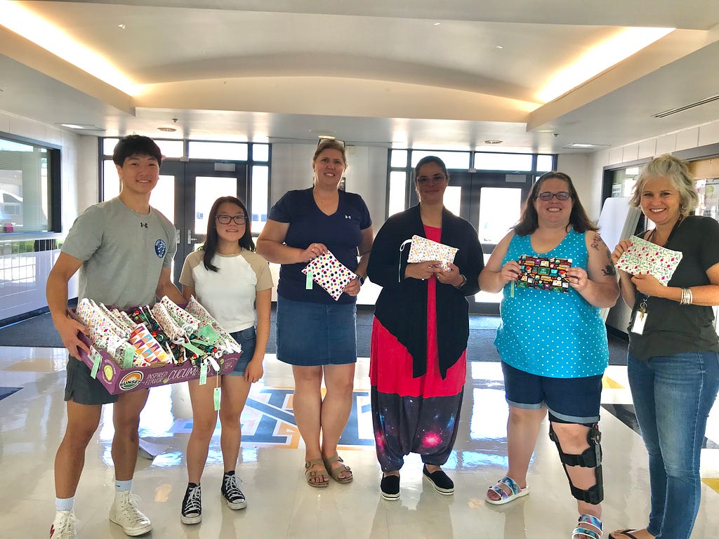Two young people and six adults standing in a school entrance. The young people are distributing mask kits to the four teachers who are smiling and displaying their colorful kits.