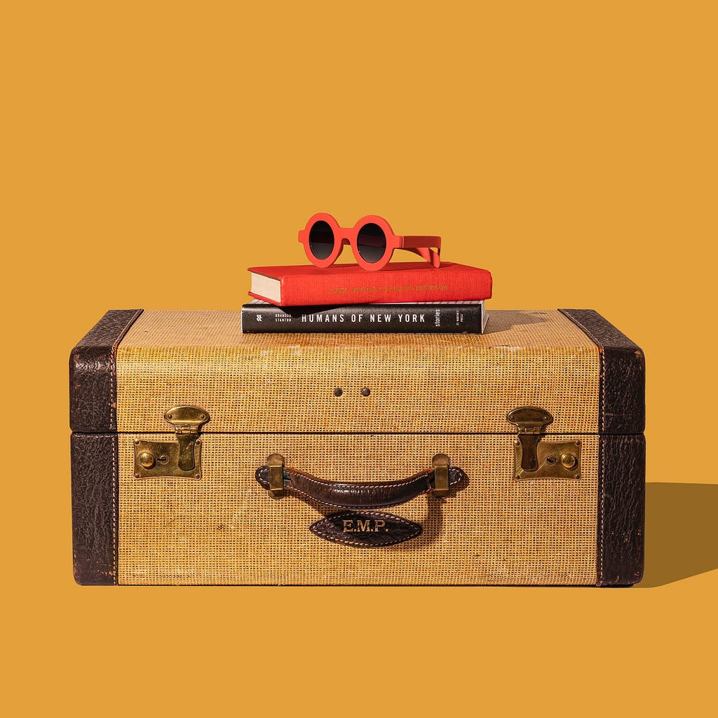 Vintage style rectangular brown suitcase with two books and round red framed sunglasses sitting on top, with orange background