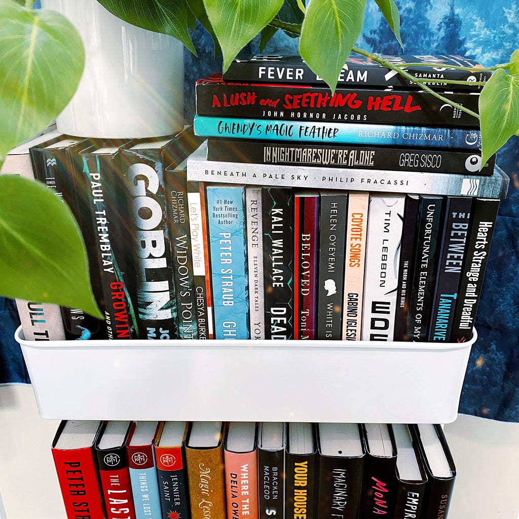 A white cart with books in it that are mostly horror titles with a plant on top.