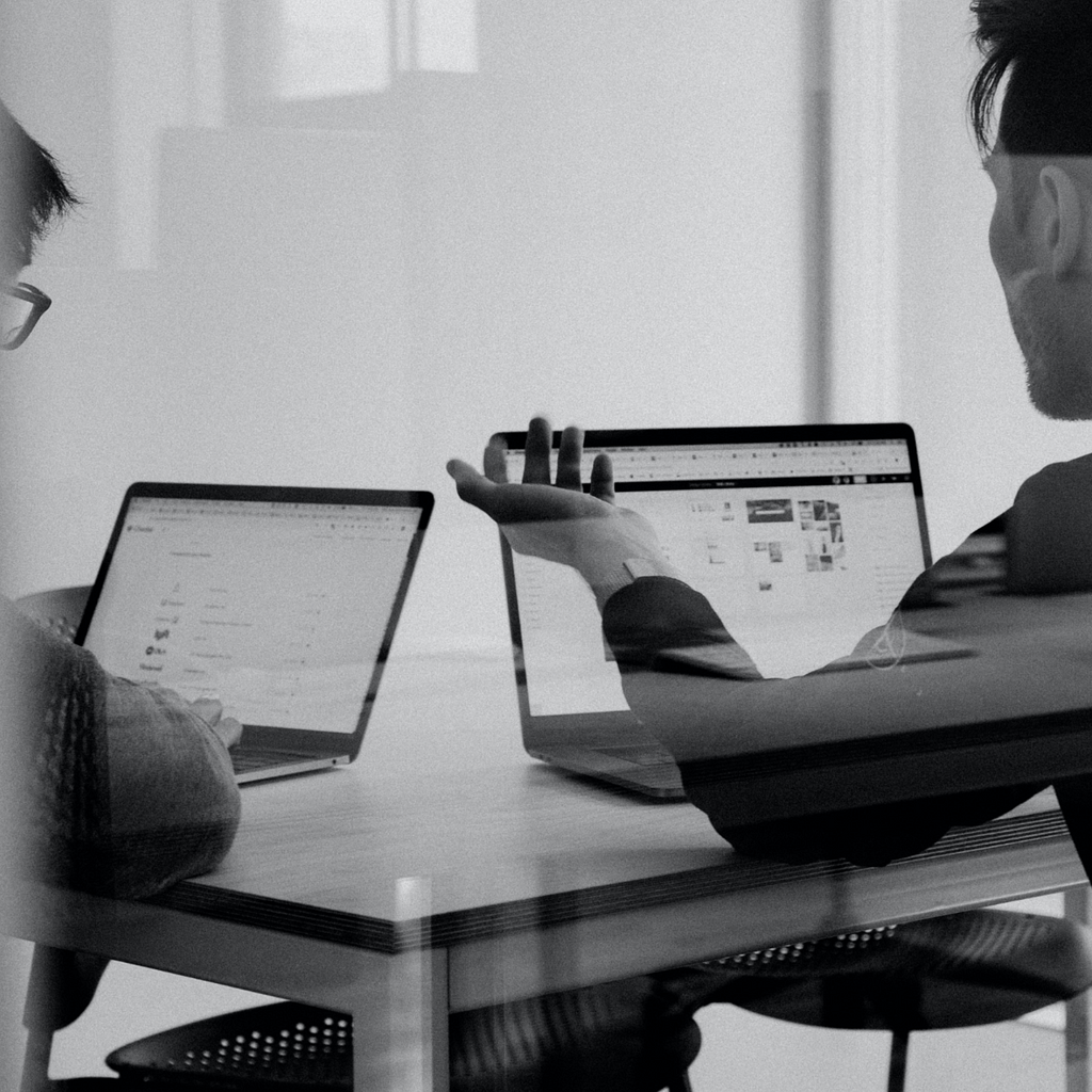 A black and white close-up photo showing two men, behind a glass window, looking and working on their laptops. They seem to be discussing work.