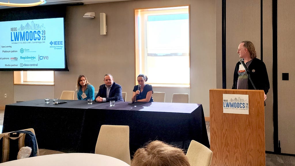 Photo of four adults in business attire. Three are sitting at a table to speak on a panel. One is standing at a podium to moderate. A screen displaying the Learning with MOOCs conference 2023 logo hangs on the wall behind them.