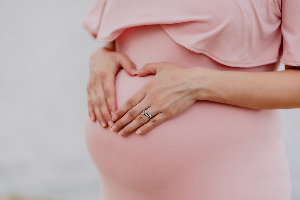 The midsection of a pregnant woman who is making a heart shape over her belly with her hands.