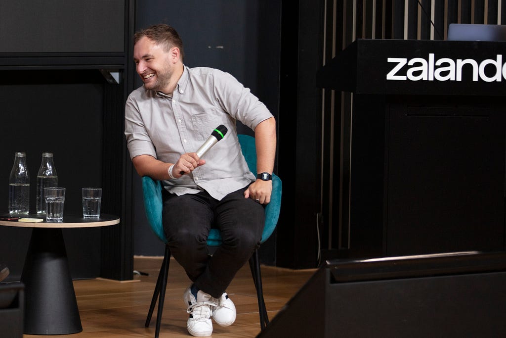 A person seated in a chair and holding a microphone, leaning forward and smiling broadly.