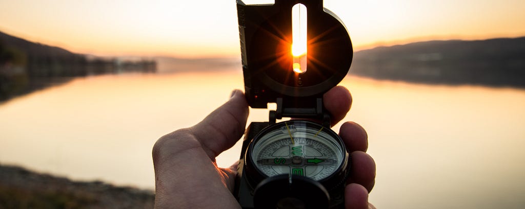 Picture of a hand holding a compass. There’s a lake in the background