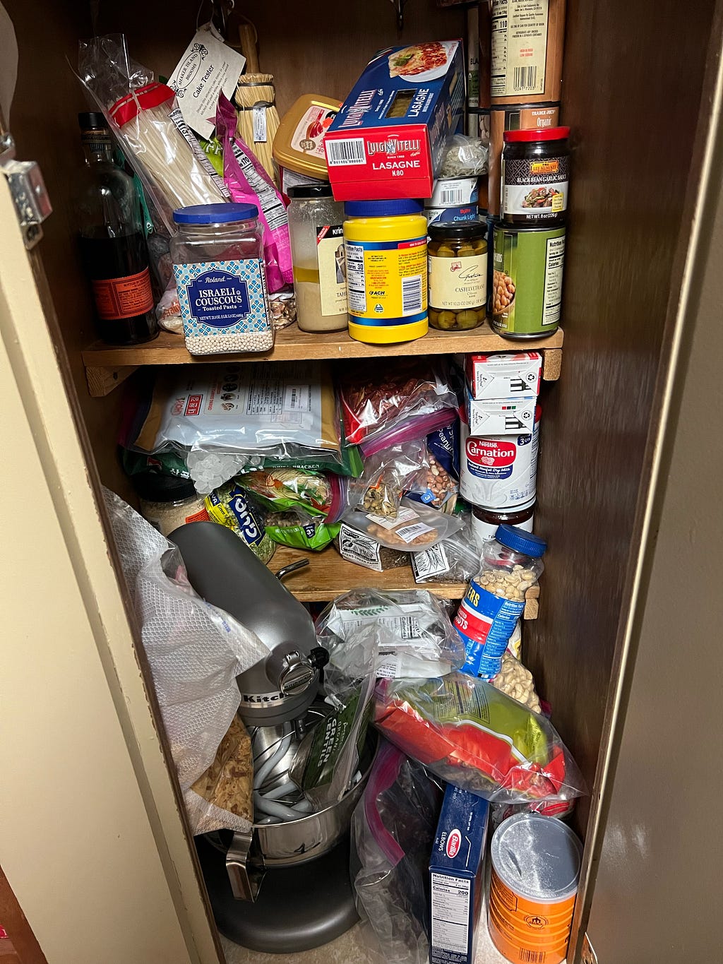 The Author’s terribly disorganized pantry cabinet containing a kitchenaid mixer and a hodgepodge of random shelf-stable ingredients.