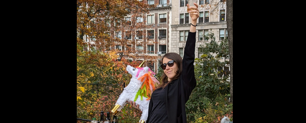 photo of Natalya holding a unicorn pinata in one hand and cheering with a glass of champagne in the other outside in central park