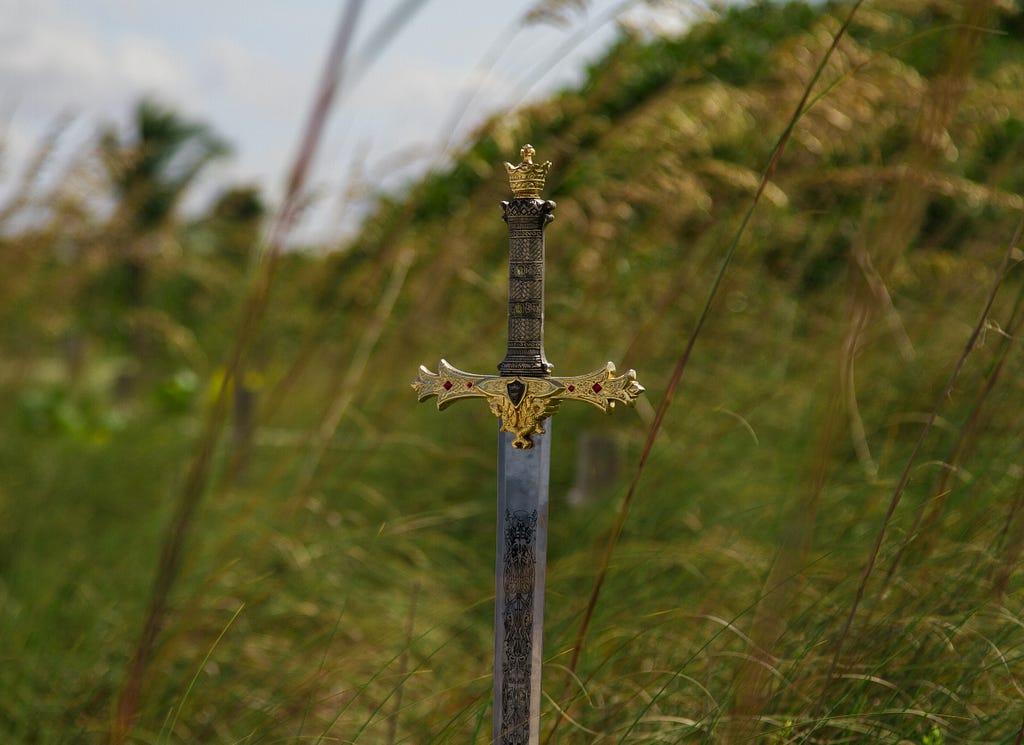 An ornate sword is stuck into the ground with a grassy field in the background.