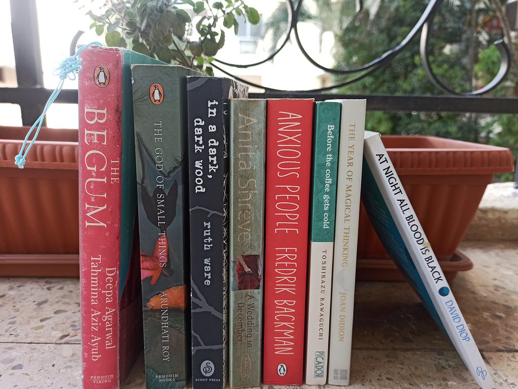 8 books are lined up in a standing position. From left to right, the books are: The Begum, The God of Small Things, In a Dark Dark Wood, A Wedding in December, Anxious People, Before the Coffee Gets Cold, The Year of Magical Thinking, At Night All Blood is Black.