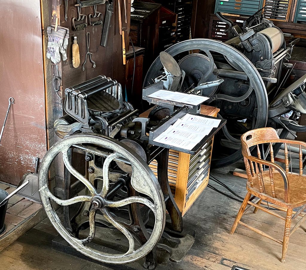 “The Liberator” foot powered press. There is a large wheel, pedals, and rollers in the device. There are sheets of paper on top. In the background is another kind of press and to the right is a chair.