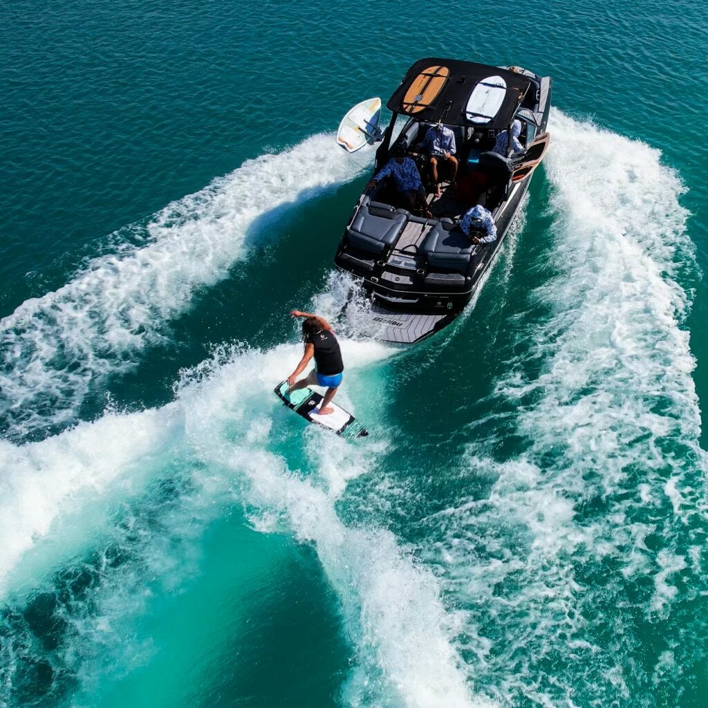 Young man wakesurfing on the lake behind a grey boat with friends.