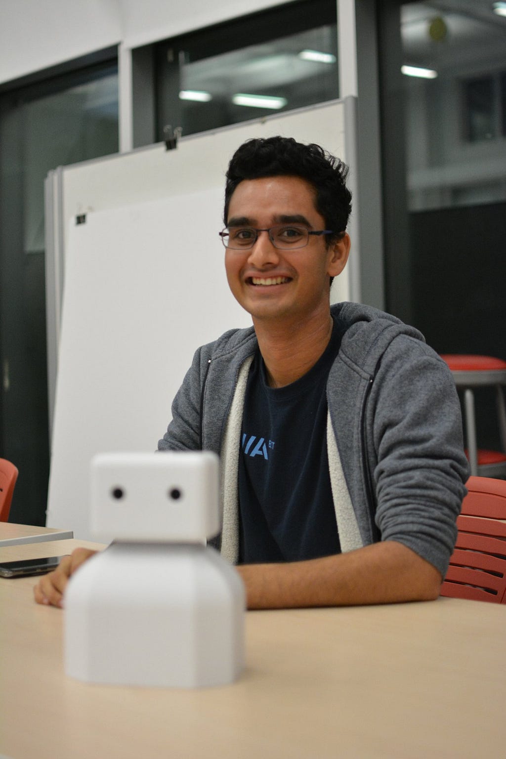 Akhil sits at a table smiling, in front of him is an out of focus robot model.