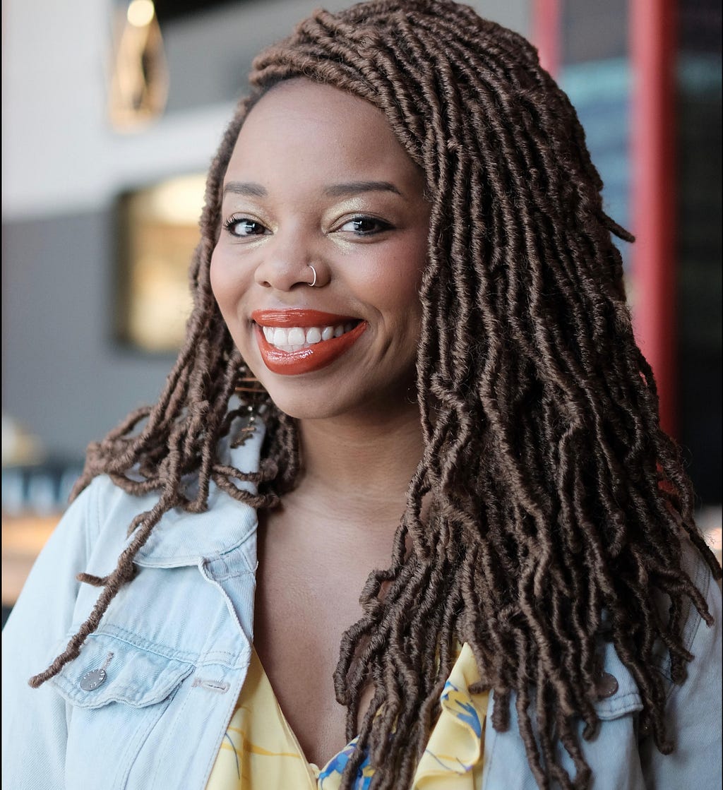 Picture of a Black woman smiling. She has braids and is wearing peach lipstick, a yellow dress, and a jean jacket.
