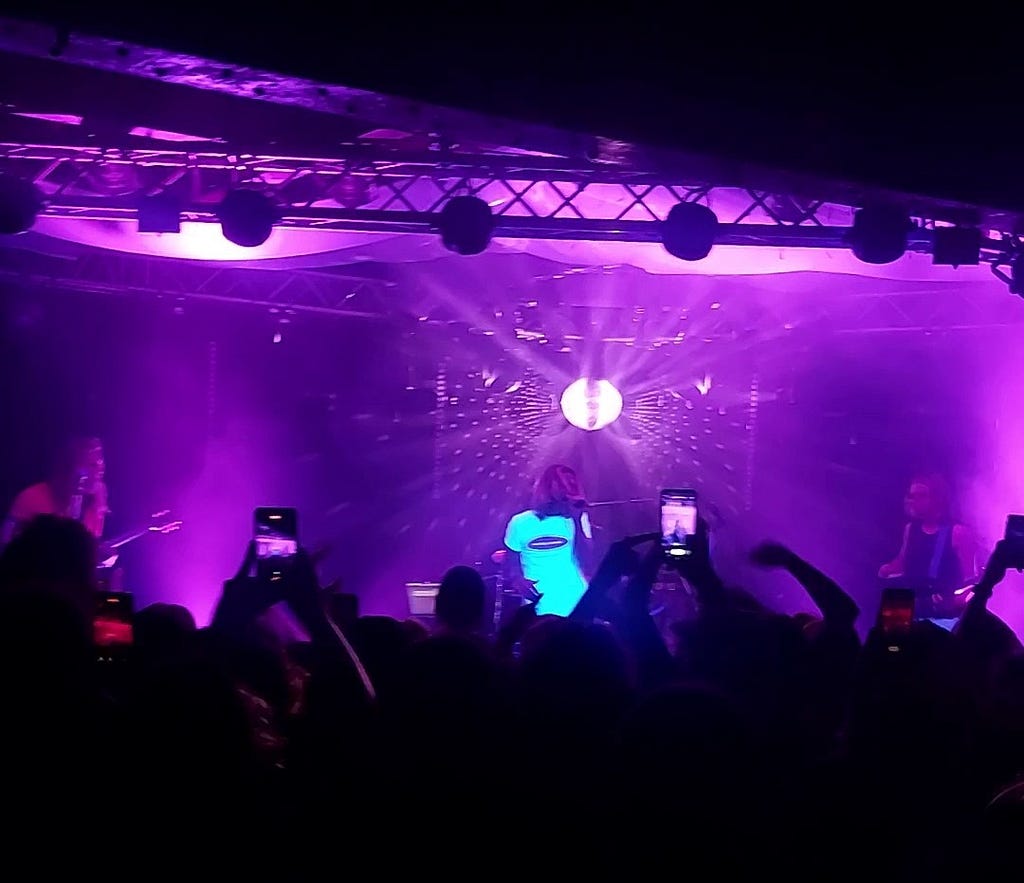 Singer in a white T-shirt, below a glitterball, bathed in purple light, on stage in front of a crowd, with hands in the air.