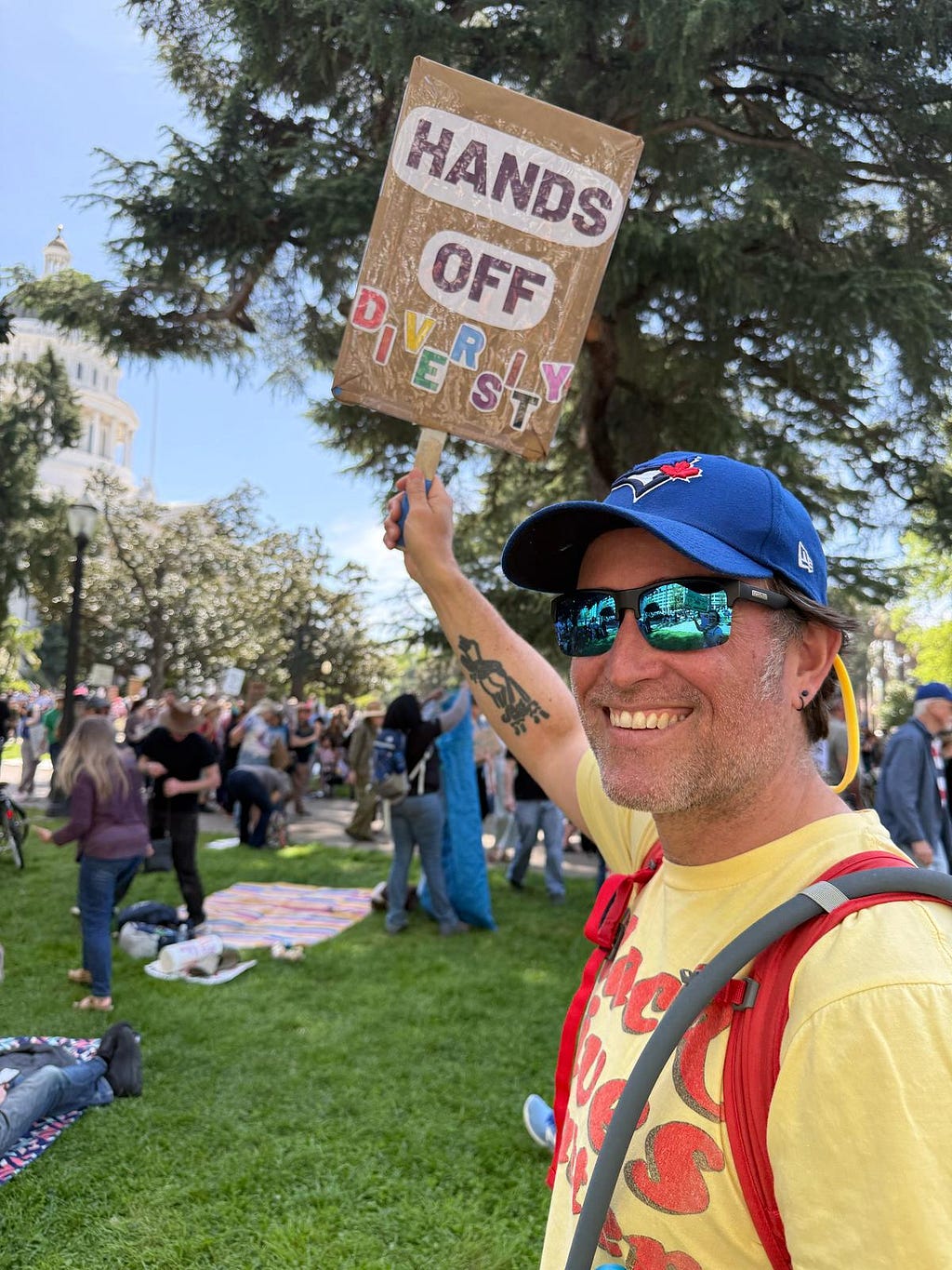 Me, wearing a Blue Jays ballcap, a yellow “Black Lives Matter” t-shirt a red backpack, and sunglasses, holding a sign that says “Hands off diversity”