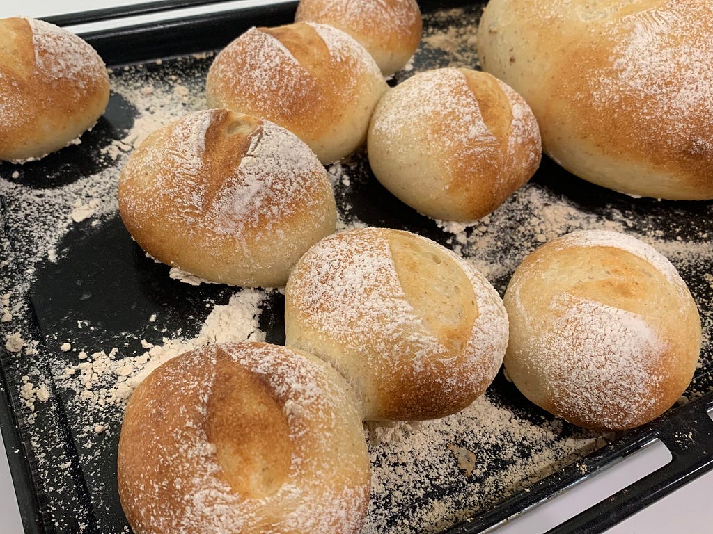 Freshly baked bread rolls and a loaf on a baking tray without an oven sheet, showing no sticking or burning. The tray is lightly dusted with flour.