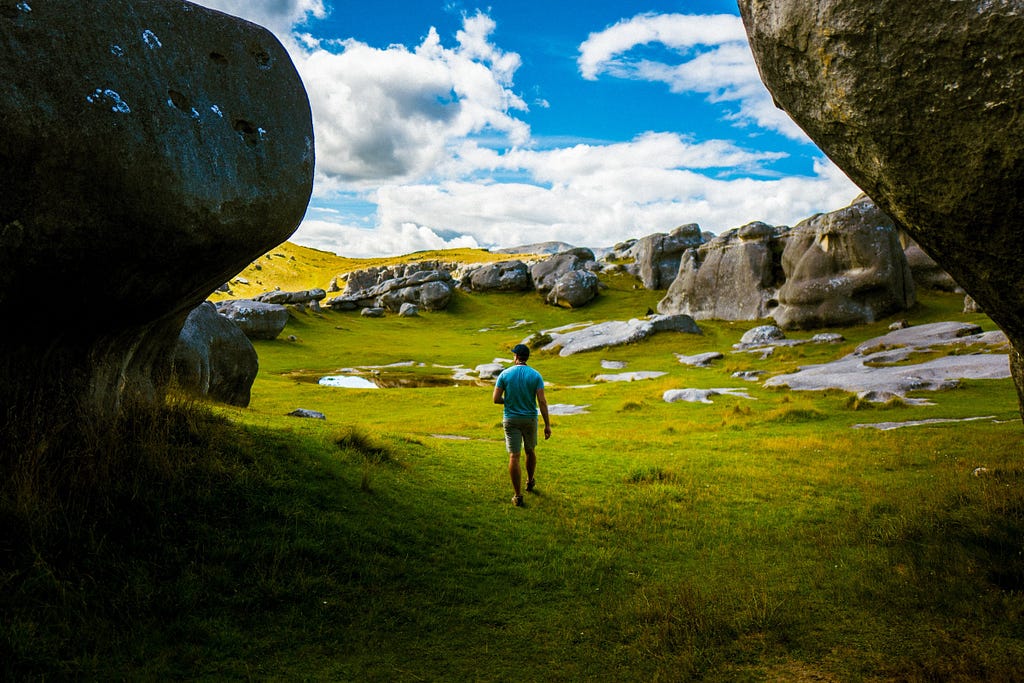 Photo of a man walking into a beautiful field in New Zealand. There are rock formations, green grass, and moss. The sky is blue but cloudy.