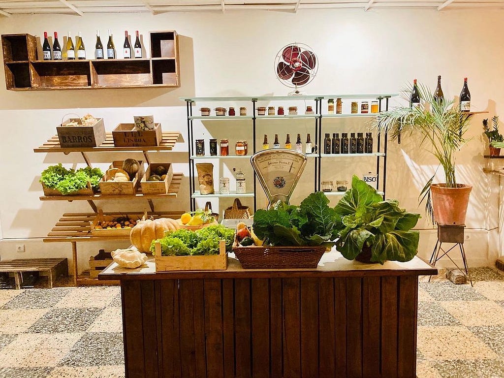 A local greengrocery cozy store with vegetables such as lettuce and tomatoes in the front area and in the back there are wine bottles, a vase with a plant, and other grocery products.