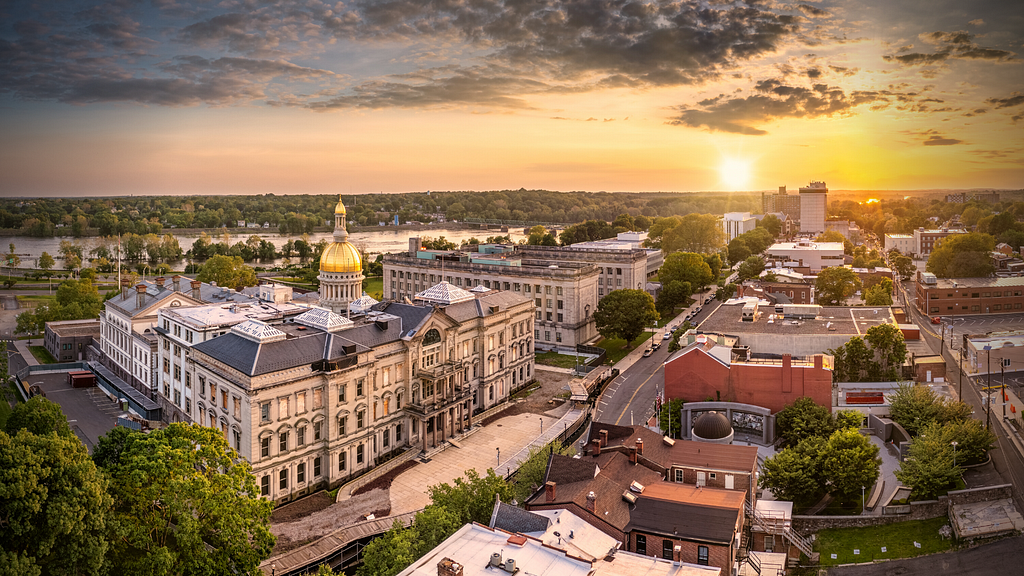 A dramatic photo of the city of Trenton at sunset, with the Trenton Statehouse in the foreground and the setting sun just about to disappear over the horizon on the right side of the photo.