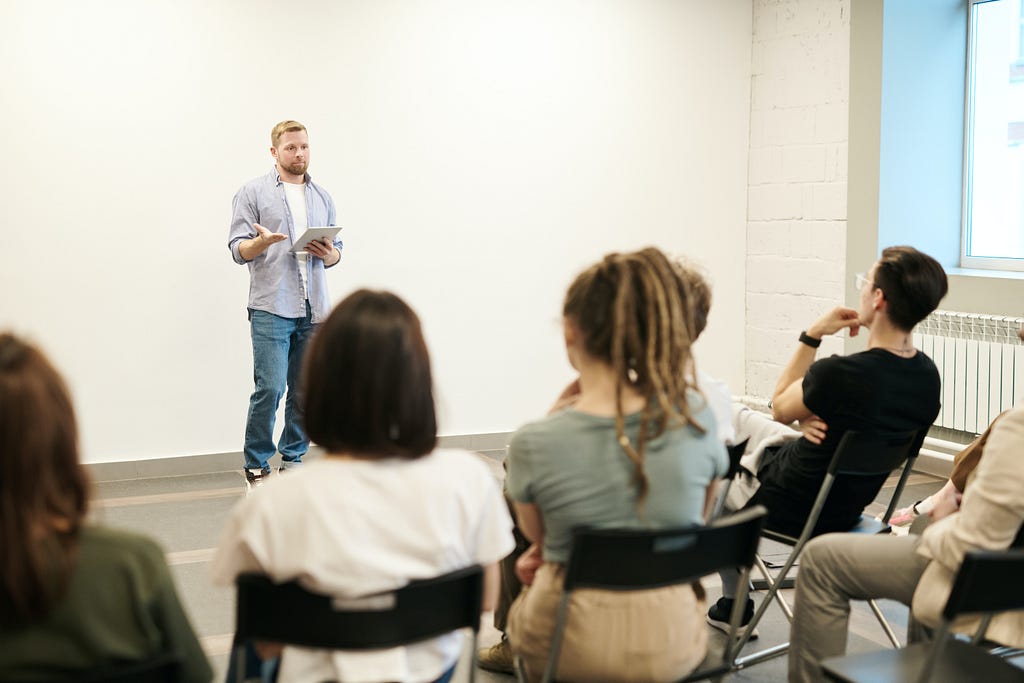 A man with blond hair holding an eye pad in his hand is speaking confidently in front of both a male and female audience sitting on chairs. The man is wearing a white tea shirt, blue jeans, and a light blue shirt.