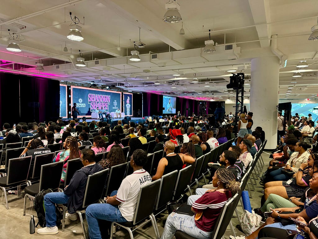 A packed auditorium of attendees listening to a keynote panel discussion with Shannon Sharpe.