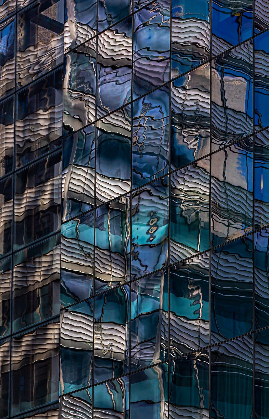 “Blue and white concrete building”, photograph by Robert Ullman (source: https://artist.scop.io/image/blue-and-white-concrete-building-73) — a building façade of square glass panes reflecting a distorted image of a concrete building in shades of dark blue, aqua, and off-white.