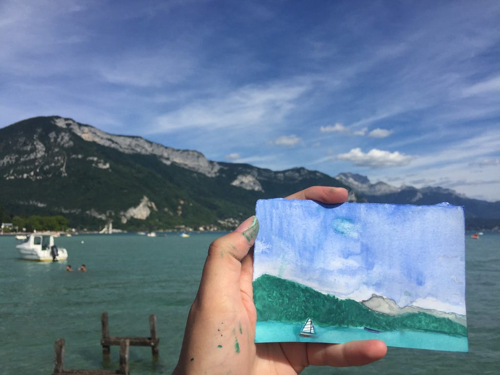 A small watercolor painting held up in front of Lake Annecy, French Alps.
