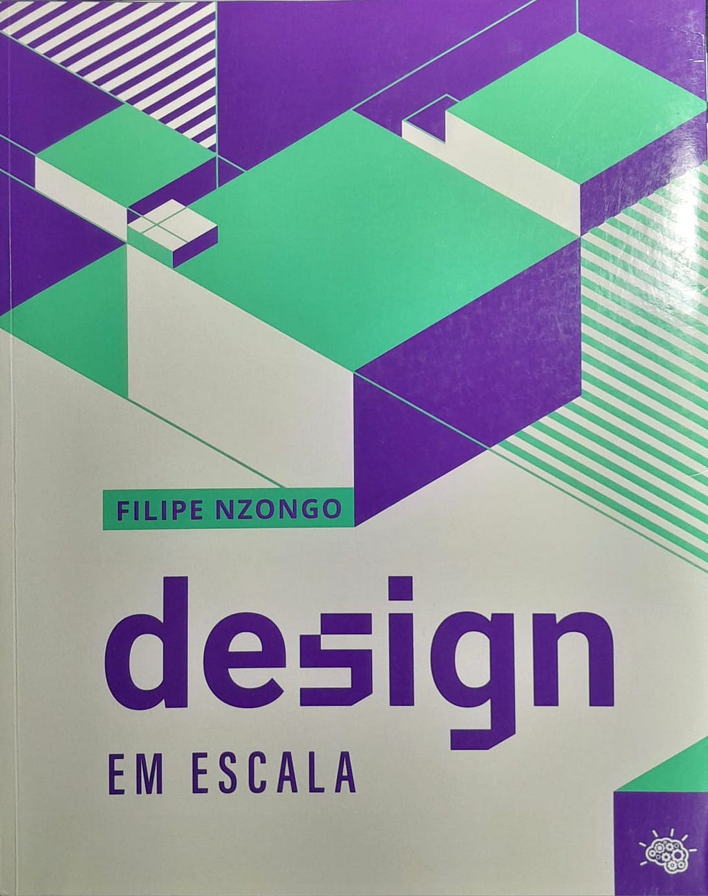 Capa do livro Design em Escala. O fundo é bege, com formas triângulares em verde, roxo e bege. Alguns triângulos possuem listras. Na parte de baixo está escrito Filipe Nzongo em roxo, dentro de um retângulo verde, “Design”, com uma fonte personalizada, e “em escala”, em caixa alta, ambos em roxo. No canto inferior direito está o logotipo da editora Viver Cultural.