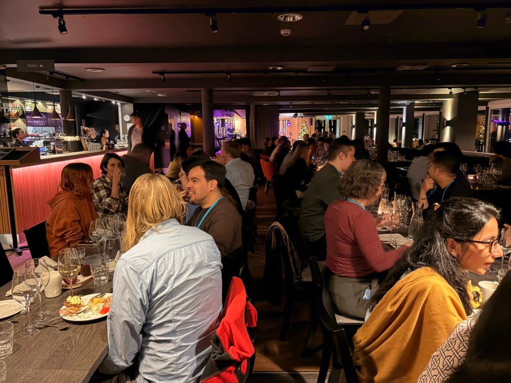 A dim room of tables, wine glasses, and a community of people talking and smiling.