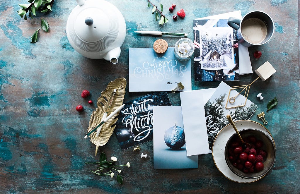 An aerial view of a writing desk, with a pen, a feather, several holiday cards, a teapot and cup of chocolate and a bowl of berries all arranged together.