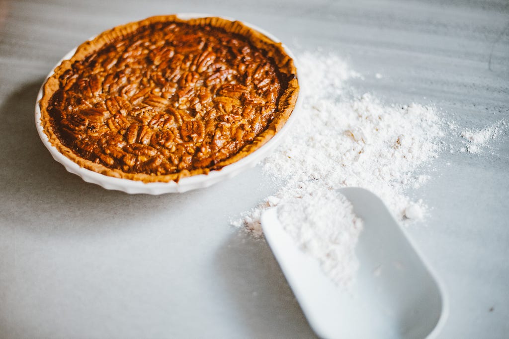 Pecan pie on a light stone background. A white plastic flour scoop with scattered baking flour is displayed next to the pie.
