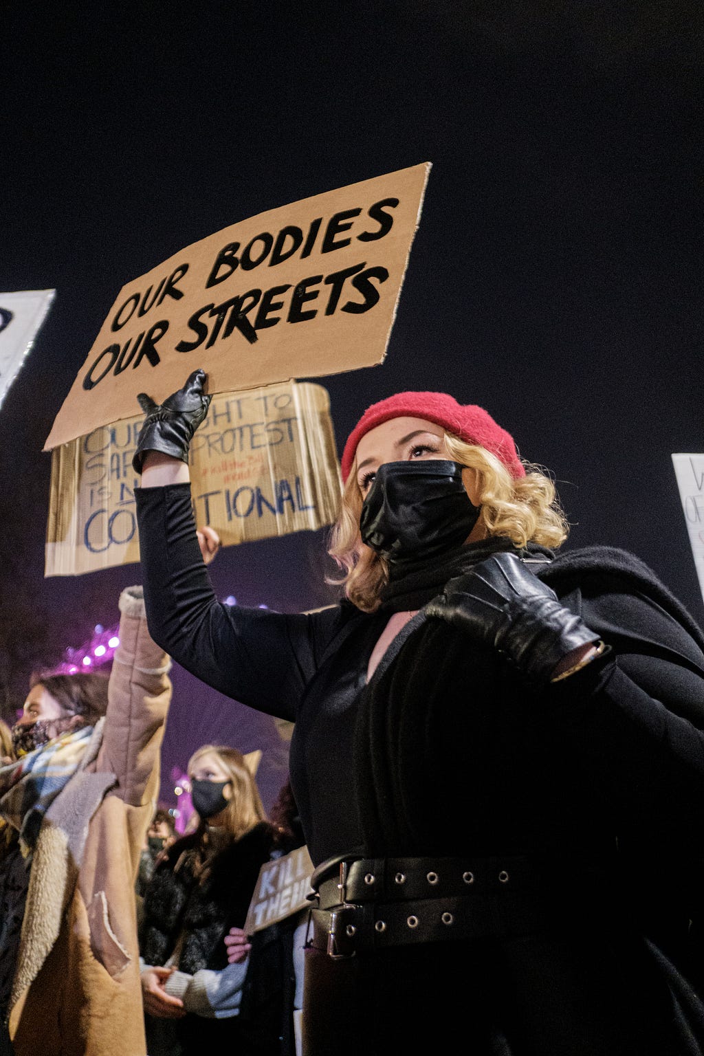 Several women protesters standing in the street, one blonde woman with a black mask holding a sign saying, “Our Bodies, Our Streets.”