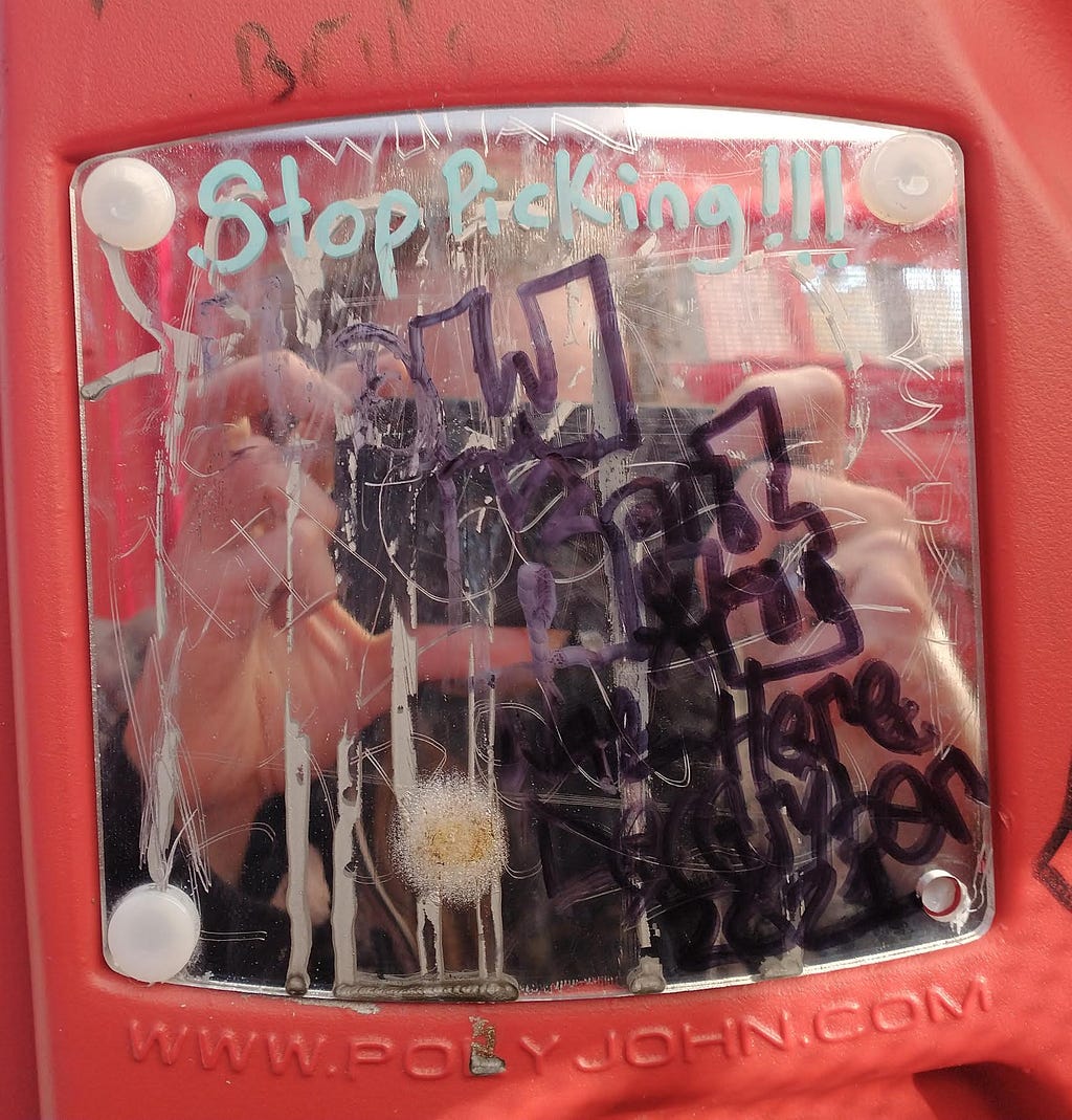 Closeup of a tiny little mirror in a porta potty. The reflection of me taking the photo on my phone is barely visible behind the graffiti. Across the top of the mirror it reads, “Stop picking!!!”