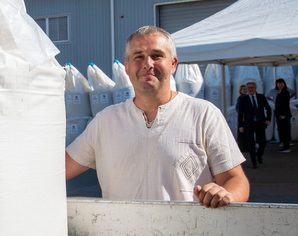 A man wearing a traditional Ukrainian shirt stands in front of bags of chemical fertilizer.