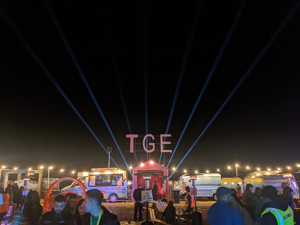Neon sign with letters TGE with strobe lighting behind them and a few food trucks in the foreground